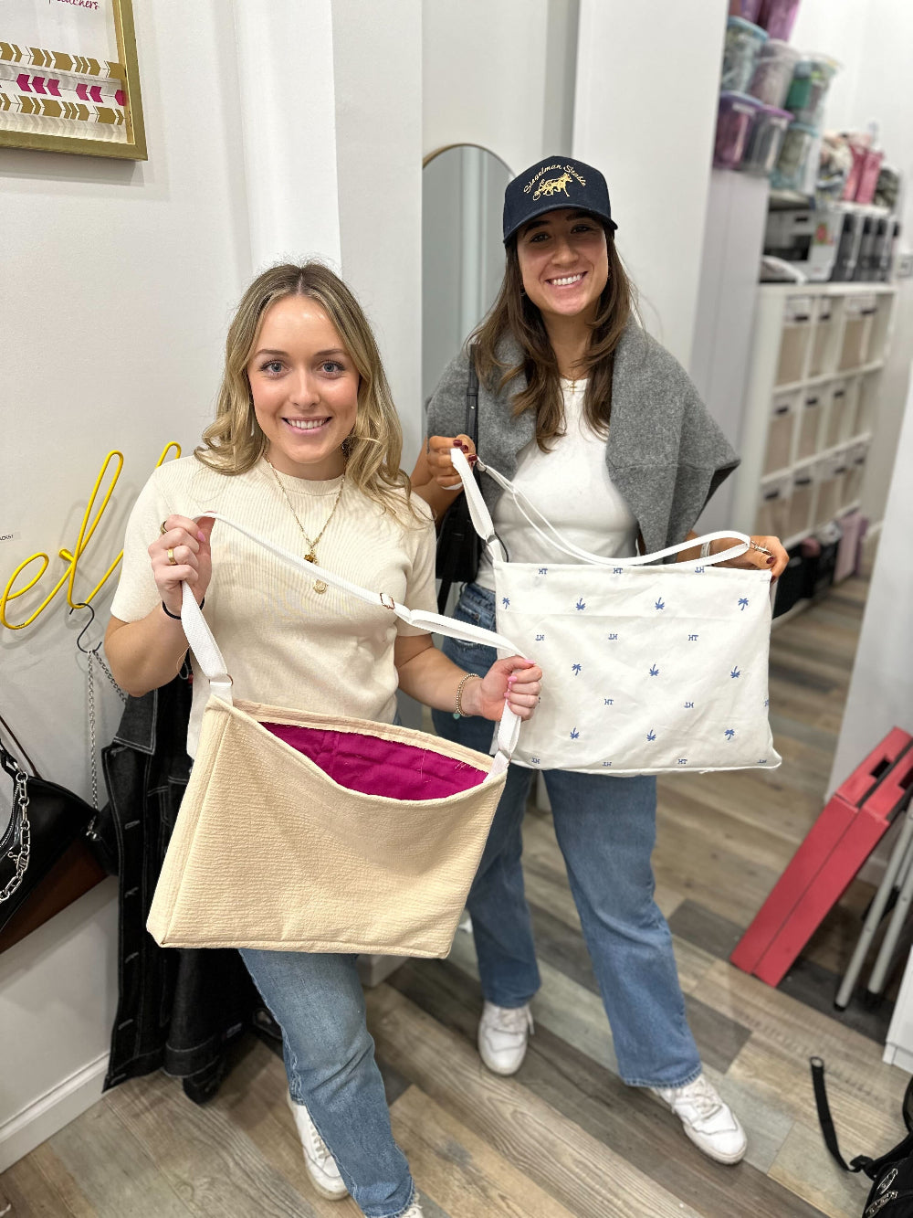 Two women standing in a boutique holding canvas tote bags. The woman in front holds a beige textured tote bag with a bright pink lining, while the woman behind her holds a white canvas tote bag with small navy blue embroidered stars.